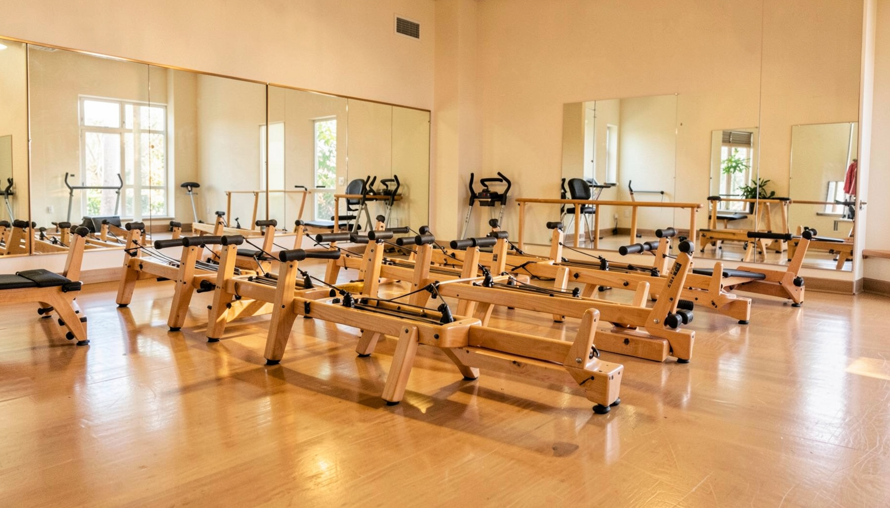 Open studio space with wooden exercise reformers lined up on a shiny floor, mirrors along the wall, and warm natural tones creating a calm clinical atmosphere