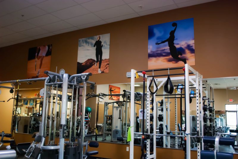 Bright gym interior with weight racks, pull-up bars, and mirrors reflecting a spacious training area with action-sports photographs on warm brown walls