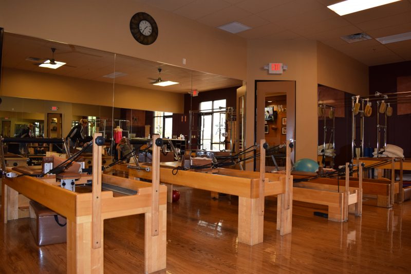 Bright studio room with wooden Pilates reformer machines lined up on a polished wood floor, mirrors along the wall, and a round wall clock